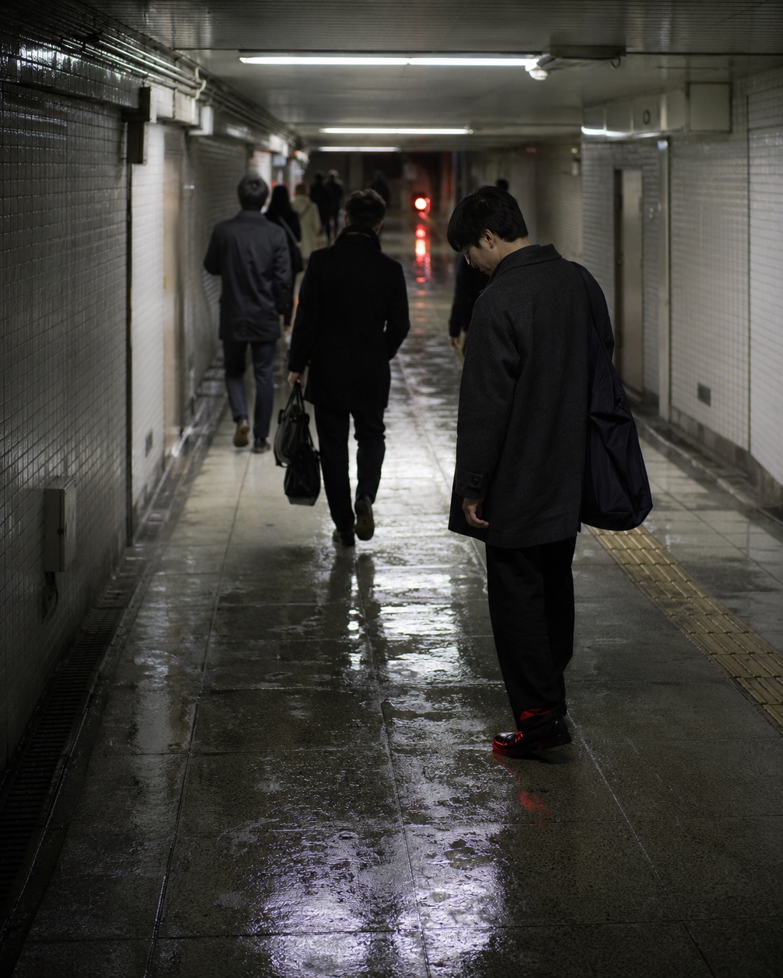 Tokyo: a man pauses in a wet underpass while commuters keep walking, a faint red reflection on his shoe